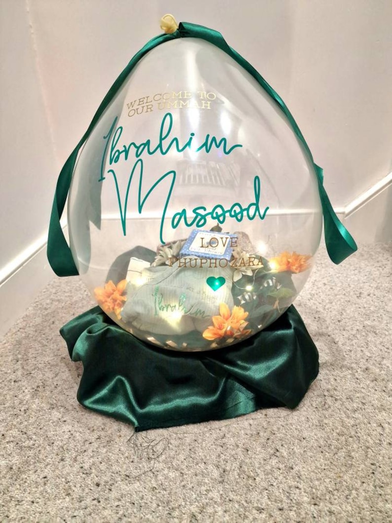 May include: A clear balloon with a green ribbon and a green satin base. The balloon has the text "Welcome to our Ummah Ibrahim Masood Love HuphOzara" written on it.