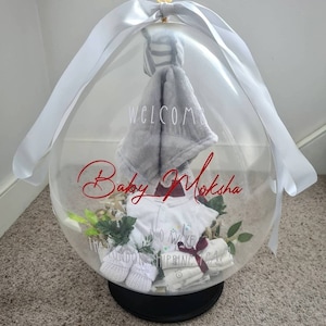 May include: A clear balloon filled with baby clothes and a blanket. The balloon is tied with a white ribbon and has the words "Welcome Baby Moksha" written on it.