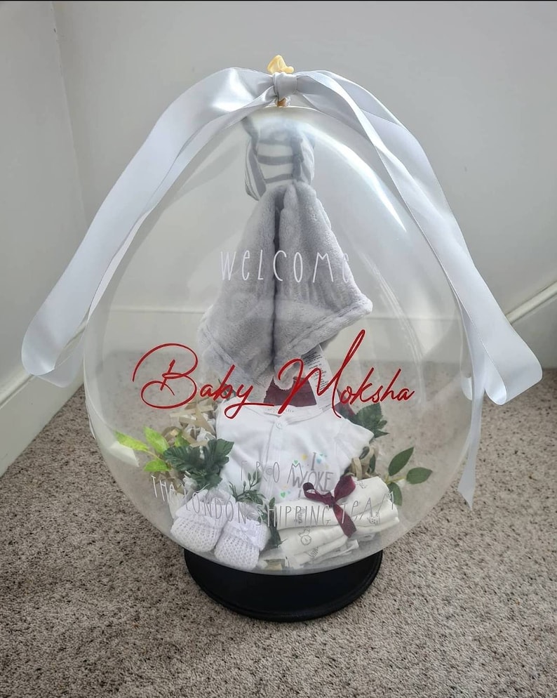 May include: A clear balloon filled with baby clothes and a blanket. The balloon is tied with a white ribbon and has the words "Welcome Baby Moksha" written on it.
