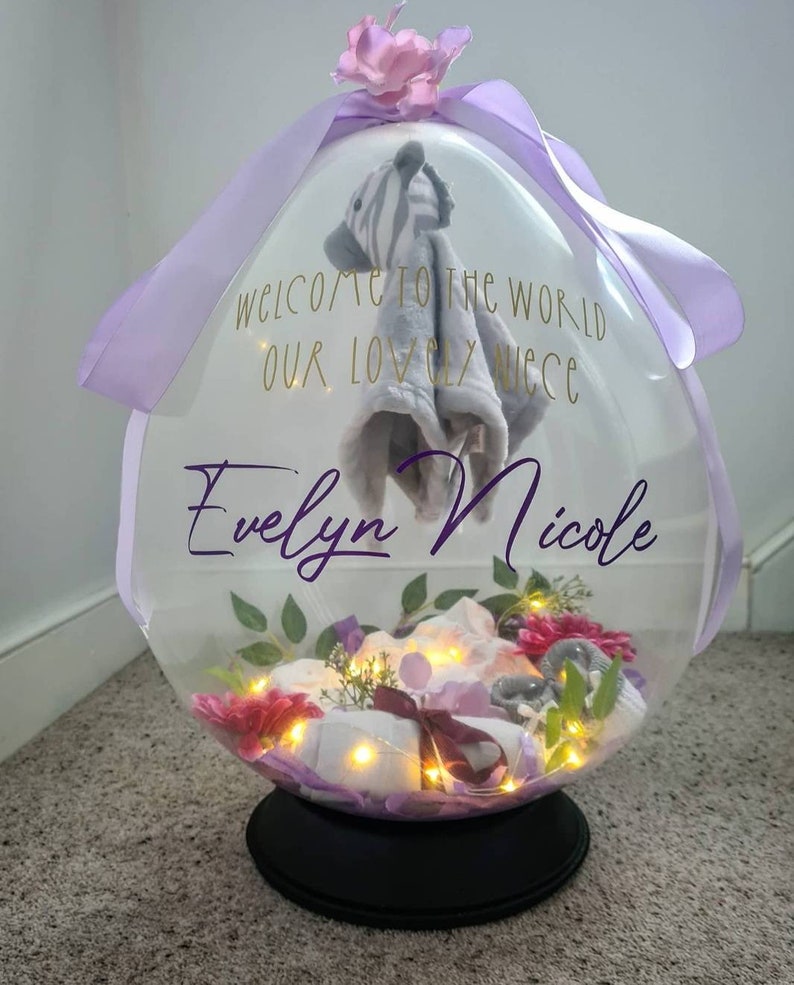 May include: A clear balloon filled with a variety of baby items, including a stuffed animal, a blanket, and flowers. The balloon is decorated with a purple ribbon and the text "Welcome to the world our lovely niece Evelyn Nicole".