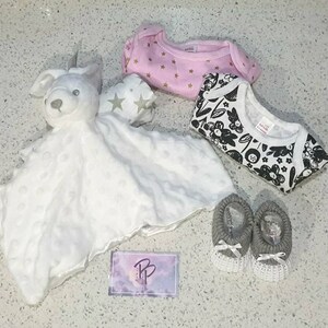 May include: A white plush bunny security blanket with a pink and gold star print onesie, a black and white floral print onesie, and two gray knitted baby booties.