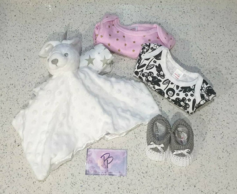 May include: A white plush bunny security blanket with a pink and gold star print onesie, a black and white floral print onesie, and two gray knitted baby booties.