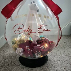 May include: A clear balloon filled with baby clothes, a stuffed animal, and lights. The balloon is tied with a red ribbon and has the text "Welcome to the world Baby Eman" written on it.