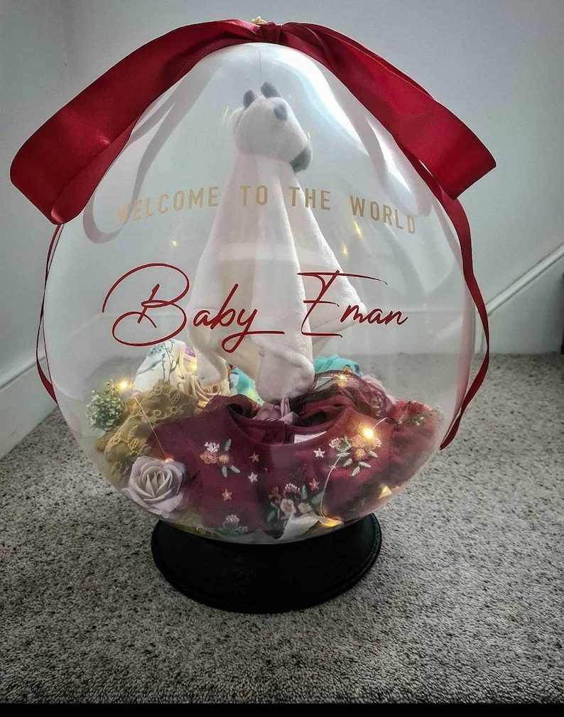 May include: A clear balloon filled with baby clothes, a stuffed animal, and lights. The balloon is tied with a red ribbon and has the text "Welcome to the world Baby Eman" written on it.