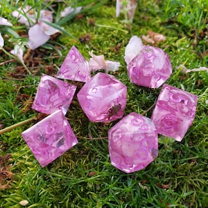 May include: Seven pink dice with white numbers on a bed of green moss. The dice are translucent and have a speckled appearance.