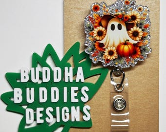 May include: A white ghost with a witch's hat and a pumpkin in a fall floral wreath with glitter accents. The wreath is attached to a clear plastic retractable badge holder with a silver clip. The badge holder is on a brown card with the text "BUDDHA BUDDIES DESIGNS" on a green leaf.
