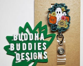 May include: A teal blue glitter badge reel with a white ghost surrounded by orange, yellow, and pink flowers. The badge reel is attached to a clear plastic retractable cord and a silver clip. The badge reel is displayed on a brown card with the text "BUDDHA BUDDIES DESIGNS" on a green leaf cutout.