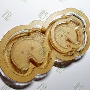 May include: Two clear acrylic cookie cutters in the shape of a cartoon bear with a heart on its chest. The cutters are layered on top of each other, with the smaller cutter inside the larger cutter. The cutters are on a brown wooden surface. The text "BUDDHA BUDDIES DESIGNS" is printed on the surface.