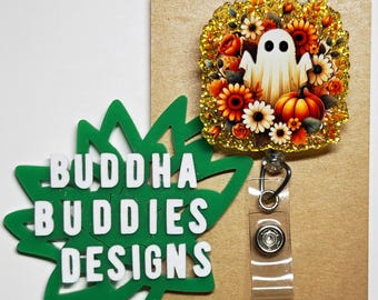 May include: A gold glitter and floral wreath with a white ghost and orange pumpkins. The wreath is attached to a clear plastic badge holder with a silver clip. The badge holder is on a brown card with the text "BUDDHA BUDDIES DESIGNS" in white letters on a green leaf.