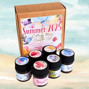 May include: A brown cardboard box set containing six small perfume oil bottles. The box is labeled "Summer 2025 Cottage Mini Sample Set." Each bottle has a black cap with a floral design and a label with a unique name. The set is based in fractional measurements.