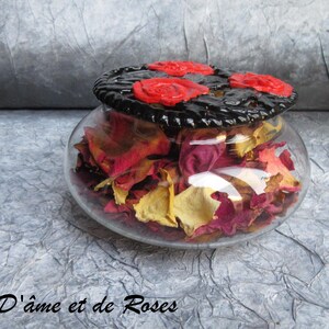 Pode incluir: Um frasco de vidro transparente cheio de pétalas de rosa secas em tons de vermelho, amarelo e rosa. O frasco tem uma tampa preta decorativa com três detalhes de rosas vermelhas. O texto "D'âme et de Roses" é visível.