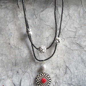Puede incluir: Un collar de cuentas negras con un dise&ntilde;o de dos hilos. El collar presenta cuentas con dise&ntilde;os en blanco y negro y un colgante con un patr&oacute;n de tablero de ajedrez y una rosa roja en el centro. El texto "D'&acirc;me et de Roses" es visible.