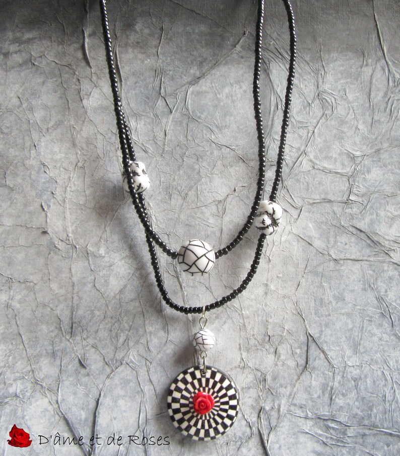 Puede incluir: Un collar de cuentas negras con un dise&ntilde;o de dos hilos. El collar presenta cuentas con dise&ntilde;os en blanco y negro y un colgante con un patr&oacute;n de tablero de ajedrez y una rosa roja en el centro. El texto "D'&acirc;me et de Roses" es visible.
