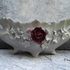 May include: A white ceramic planter with a scalloped edge and two handles. The planter is decorated with raised floral details and a single red rose in the center. The planter is sitting on a gray textured surface. The text "D'âme et de Roses" is visible in the lower right corner of the image.