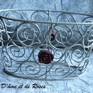 Puede incluir: Una cesta de metal plateada con un diseño decorativo de espiral. La cesta es de forma ovalada y tiene tres rosas rojas sujetas al lateral.