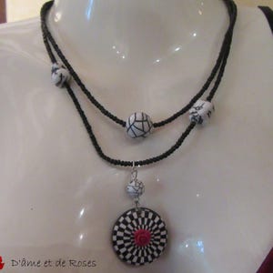 Puede incluir: Un collar de dos hilos con cuentas negras y cuentas con dise&ntilde;os en blanco y negro. El colgante presenta un dise&ntilde;o a cuadros en blanco y negro con una rosa roja en el centro. El texto "D'&acirc;me et de Roses" es visible.