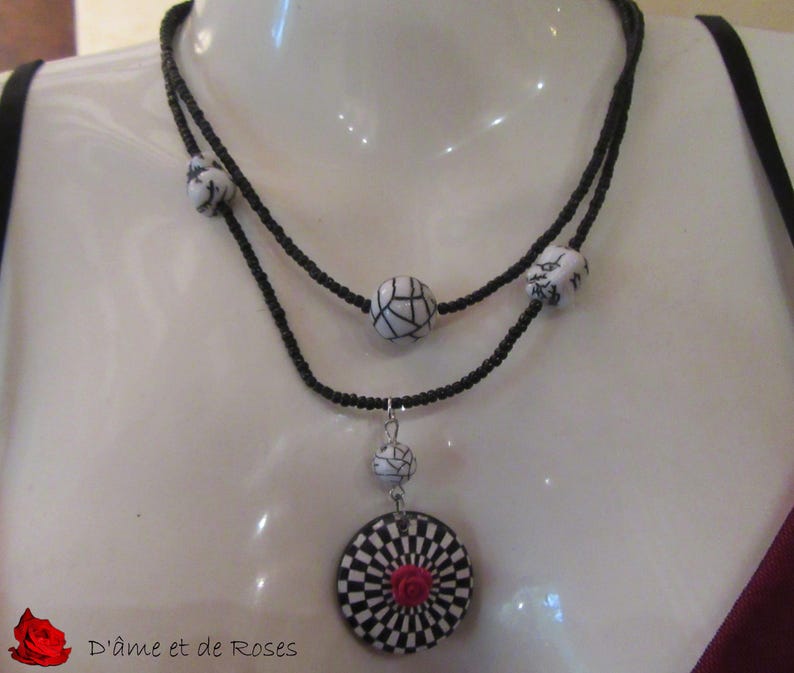 Puede incluir: Un collar de dos hilos con cuentas negras y cuentas con dise&ntilde;os en blanco y negro. El colgante presenta un dise&ntilde;o a cuadros en blanco y negro con una rosa roja en el centro. El texto "D'&acirc;me et de Roses" es visible.
