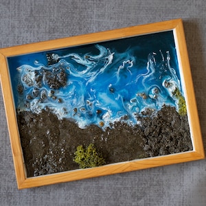May include: A framed resin art piece depicting a blue ocean with white waves crashing on a black sandy beach. The piece is framed in a natural wood frame.