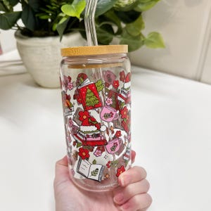 May include: A clear glass tumbler with a bamboo lid and a glass straw. The tumbler is decorated with a festive pattern of books, Christmas trees, flowers, and teacups in red, pink, and green. The tumbler is held by a person.