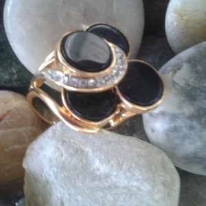 May include: A gold ring with three black stones and a row of clear stones around the largest stone.