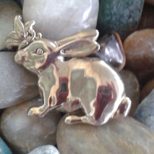 May include: A silver rabbit charm with a floral design on its head. The charm is sitting on a bed of smooth, gray stones.