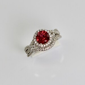 May include: A white gold engagement ring with a red ruby center stone surrounded by a halo of smaller diamonds. The ring is set with a delicate, twisted band.