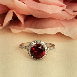Könnte beinhalten: Ein silberner Ring mit einem großen, runden, roten Edelstein, der von einem Heiligenschein aus kleineren, klaren Edelsteinen umgeben ist.