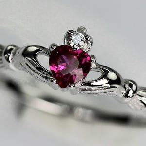 May include: A silver Claddagh ring with a heart-shaped, deep pink gemstone at the center. The ring features two hands clasping a heart, topped with a small crown and a clear, round gemstone. The ring is set against a soft, blurred background.