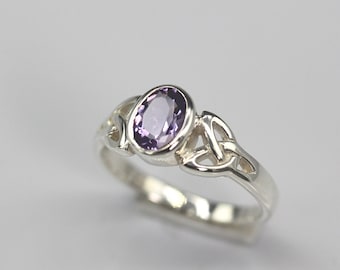 May include: A silver ring with a light purple oval gemstone. The ring has a Celtic knot design on the sides of the band. The gemstone is set in a bezel setting, and the band is smooth and polished.
