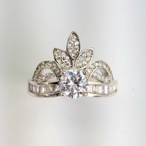 May include: A silver ring featuring a large, clear, round centre stone and a crown-like design with smaller stones. The ring is displayed against a white backdrop.