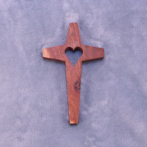May include: A wooden cross with a heart-shaped cutout in the center. The cross is a light brown color and is set against a light blue background.