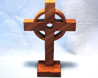 May include: A standing Celtic cross crafted from polished brown wood with a visible grain pattern. The cross features a central circular ring intersecting the vertical and horizontal bars, mounted on a rectangular wooden base for stability.