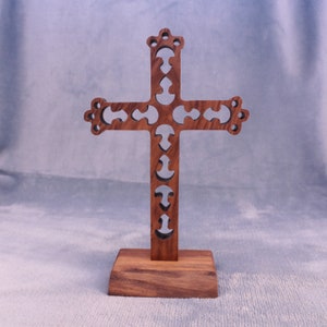 May include: A decorative wooden cross with an intricate, cut-out design and a crown-shaped top. The cross is a dark brown colour and is mounted on a rectangular base. The height is approximately 20 cm.