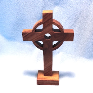 May include: A brown wooden Celtic cross featuring a central circular ring and a small hole in the middle. The cross is mounted on a rectangular wooden base, showcasing a natural wood grain pattern throughout the structure of the object.