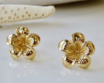 May include: A pair of gold-tone flower-shaped stud earrings. The earrings are small and delicate, and they have a textured surface.