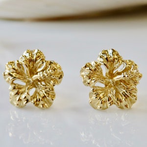 May include: A pair of gold-tone hibiscus flower stud earrings. The earrings are made of a textured metal and have a detailed design.