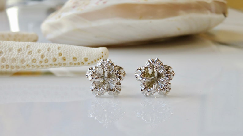 May include: A pair of silver hibiscus flower stud earrings. The earrings are small and delicate, with a detailed design.