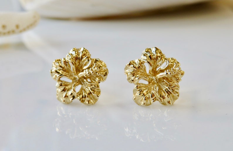 May include: A pair of gold-tone hibiscus flower stud earrings. The earrings are made of a textured metal and have a detailed design.