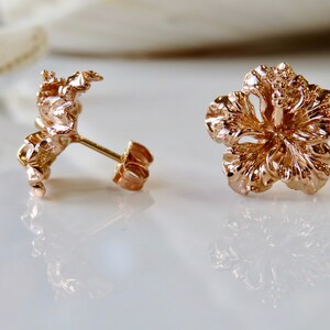 May include: A pair of rose gold hibiscus flower stud earrings. The earrings are made of a detailed, textured metal.