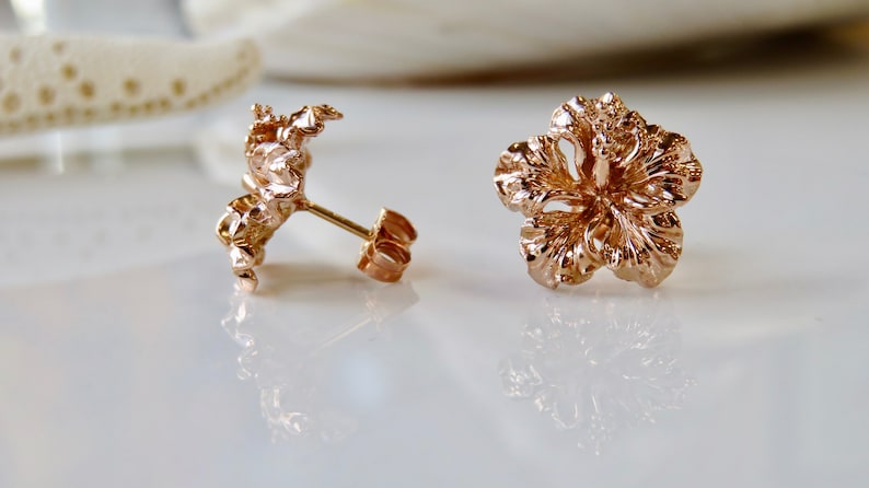 May include: A pair of rose gold hibiscus flower stud earrings. The earrings are made of a detailed, textured metal.