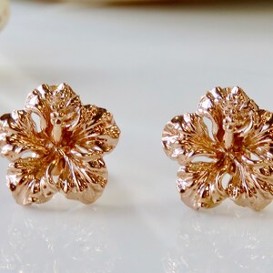 May include: A pair of rose gold hibiscus flower earrings. The earrings are small and delicate, with a detailed design.