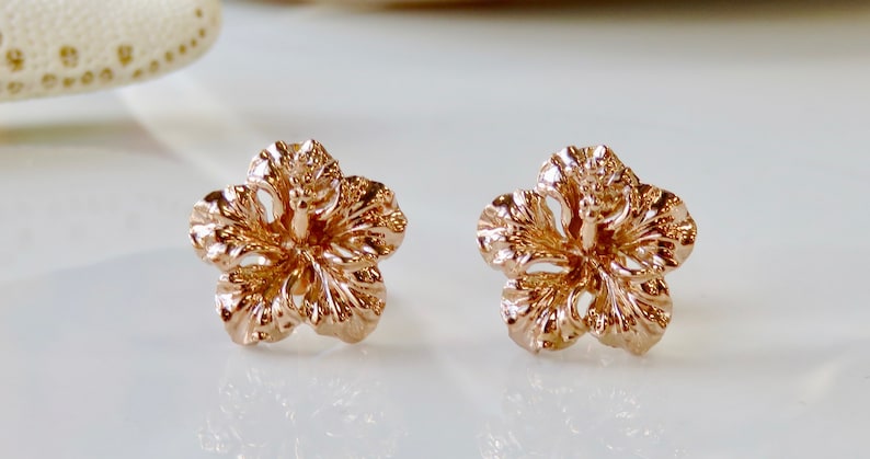 May include: A pair of rose gold hibiscus flower earrings. The earrings are small and delicate, with a detailed design.