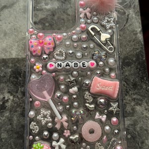 Può includere: Custodia per telefono trasparente decorata con abbellimenti rosa e argento. Presenta un pompon rosa, fiocchi, perle, un lecca-lecca e il nome "NABE". Include una fragola, una ciambella e una caramella "Sweet".