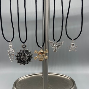 May include: Five silver and gold necklaces with different pendants hanging on a silver stand. The pendants include a skeleton, a sun, a skull and crossbones, a bird, and a cross and bones.