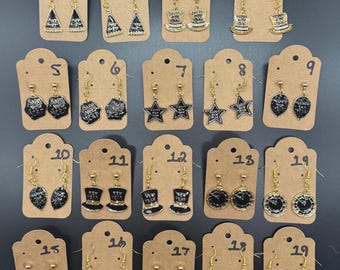 May include: A collection of nineteen pairs of gold-toned earrings on brown tags. The earrings feature various designs, including top hats, stars, clocks, and the text "Happy New Year" and "Pop Clink". The earrings are black and gold.