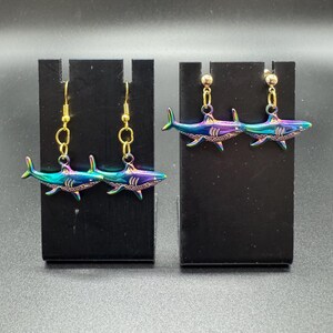 May include: A pair of shark earrings with a gold-colored hook and chain. The shark pendants are iridescent, displaying shades of blue, green, and purple. The earrings are displayed on black stands against a dark background.