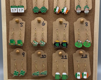 Könnte beinhalten: Zwölf Paar goldfarbene Ohrringe mit verschiedenen Motiven zum Thema St. Patrick's Day, darunter Kleeblätter, Kobolde, Bierkrüge und irische Flaggen. Die Ohrringe sind auf einer Korktafel mit nummerierten Etiketten ausgestellt.