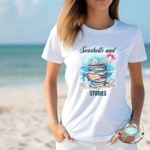 Op de afbeelding: Wit T-shirt met een aquarelontwerp van boeken, schelpen en golven. De tekst "Seashells and STORIES" is op het shirt gedrukt. Het shirt is een casual kledingstuk. Het ontwerp roept een strandthema op.