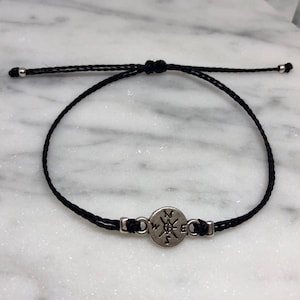 May include: Black cord bracelet featuring a silver-colored compass charm. The adjustable bracelet has a sliding knot closure and small silver beads at the ends. The compass charm has directional markings.