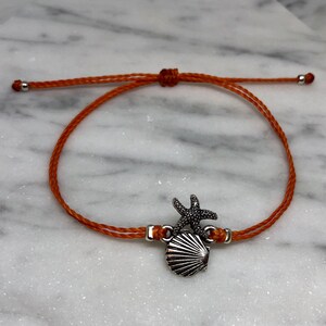 May include: Orange cord bracelet featuring a silver-colored starfish and seashell charm. The bracelet has adjustable sliding knots and silver-colored end caps. A nautical-themed accessory.